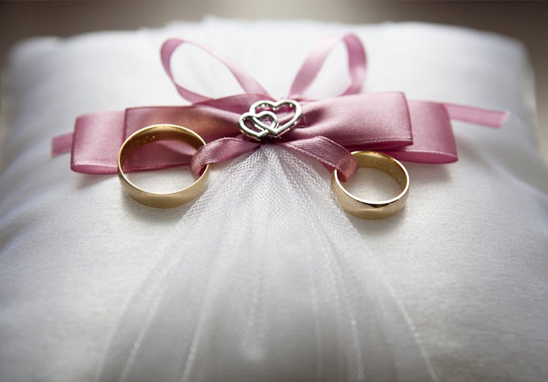 Two gold wedding rings ties to a pink ribbon on a white ring pillow