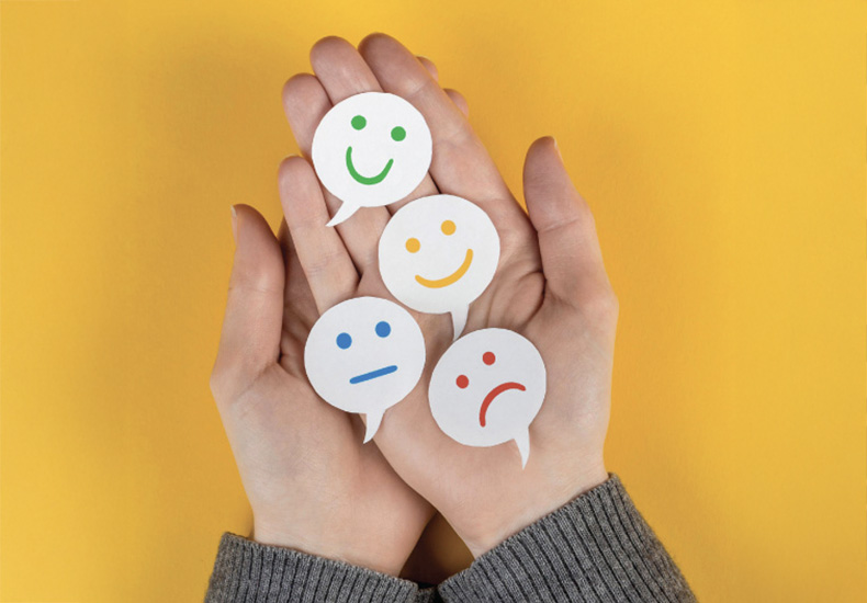 A pair of hands, palm up, holding speech bubbles cut out of paper with smiley faces, a frowning face and a neutral face drawn on them.