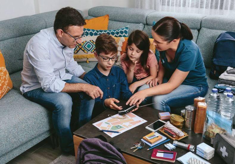 Family gathered around a coffee table with emergency preparedness kit items, including a paper plan, keys, bottled water, snacks, first-aid kit, flashlights and batteries and more.